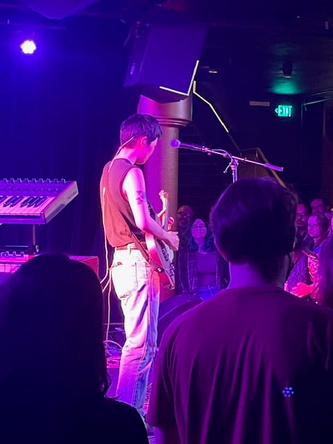 Thin Asian musician with light skin, short black hair, and in a sleeveless shirt, playing the guitar in front of a mic and a packed audience.