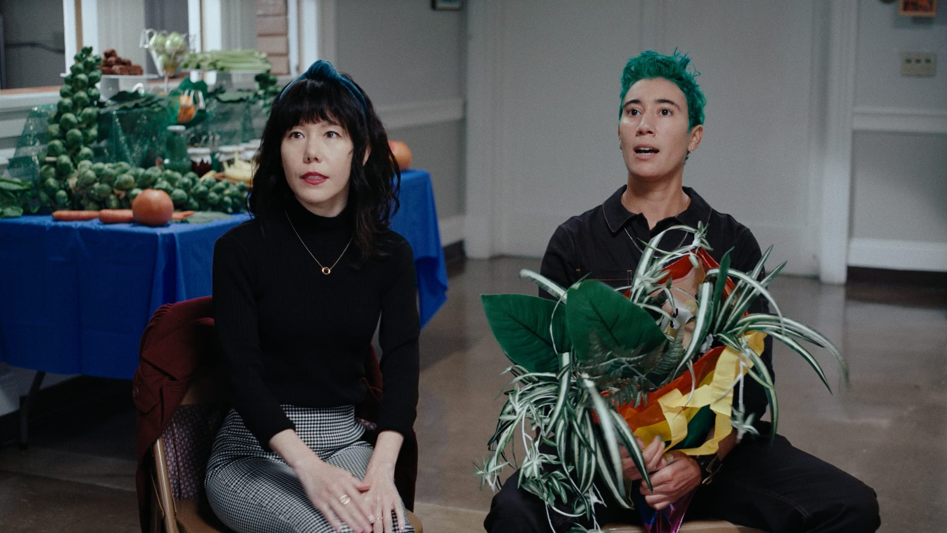 Main characters from RECKLESS SPIRITS sit next to each other, holding a bundle of plants and with a table piled with fruit in the background.