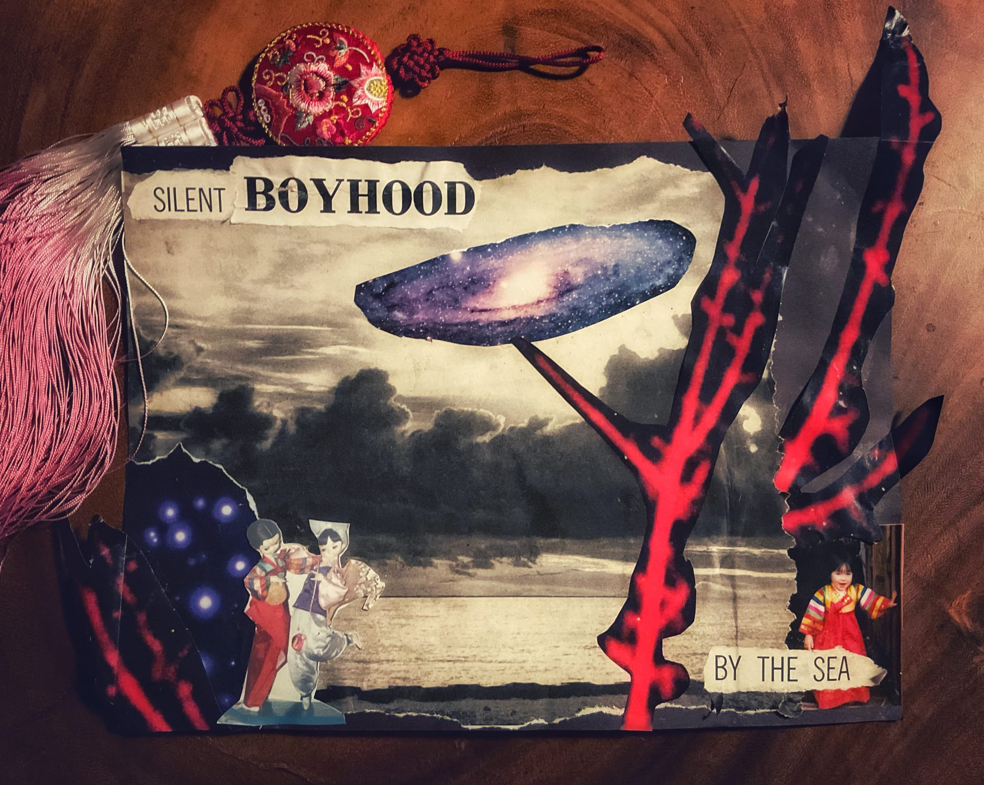 Photo of a red, white, and pink norigae (Korean silk ornament tassel traditionally worn with women's hanbok) laying next to a collage artwork on a dark wood table. The collage features a greyscale image of a late-afternoon beach surrounded by cutout images of blue stars in space, a purple-blue spiral galaxy, red coral branches, and two photographs: 1) a girl-boy pair of traditional Korean silk dolls in hanbok; 2) the artist as a child wearing a red and yellow hanbok with multi-colored sleeves. Framing the top left and bottom right, black text on white background reads: "SILENT BOYHOOD", "BY THE SEA".