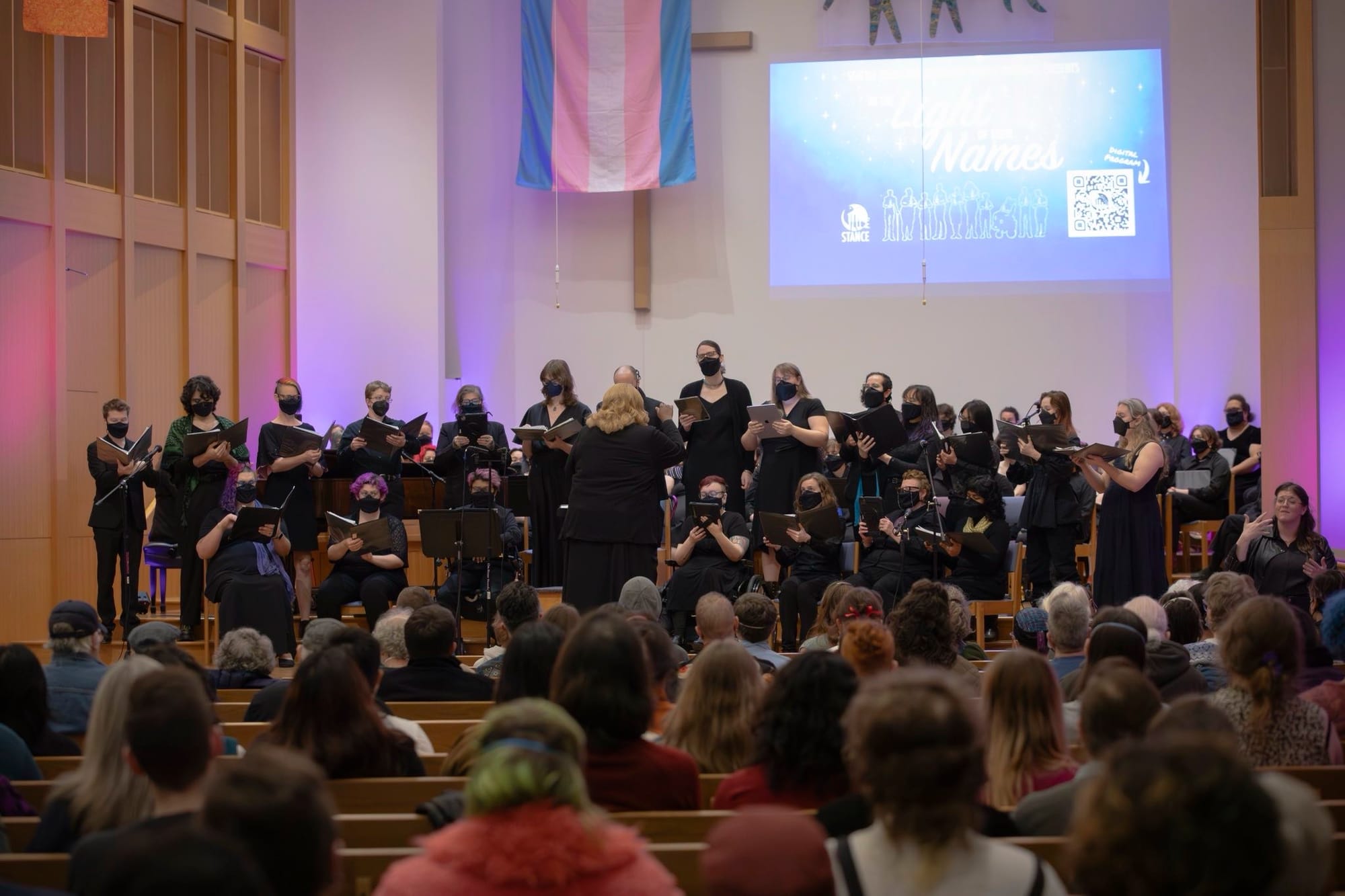 STANCE choir singing onstage at their TDoR concert with a trans flag in the background, audience members in foreground