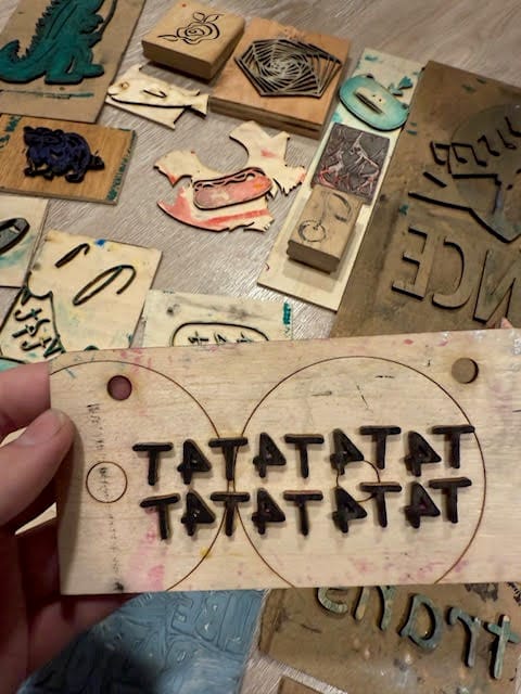 Hand holding a wood block stamp that says T4T4T4T4T4T, with many other creative stamps in the background on a table