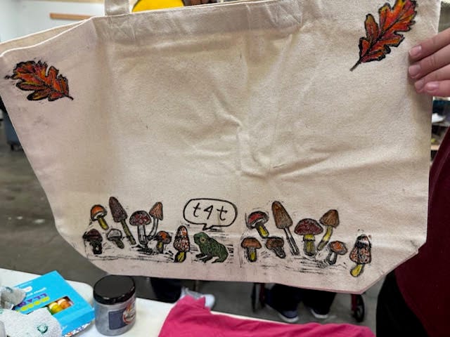 A tote bag painted with autumn leaves, mushrooms, and a frog with a speech bubble that says T4T