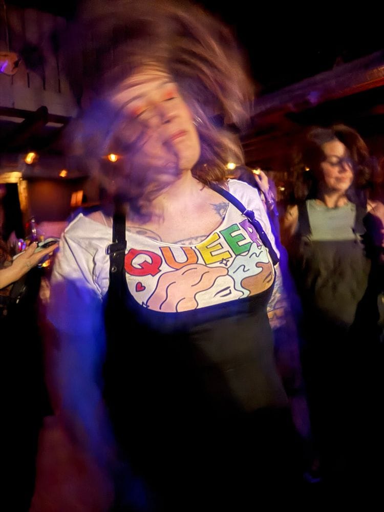 A white fem-presenting person with brown hair swooshing while dancing in a club, wearing black overalls and a white shirt that says QUEER in rainbow colors and shows two girls kissing.