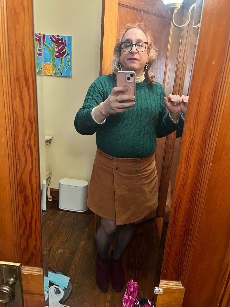 An older trans gal with glasses, light skin, and golden shoulder-length hair takes a selfie in a full length mirror, wearing a green sweater, brown corduroy wrap skirt, sheer black tights, and maroon ankle pumps.