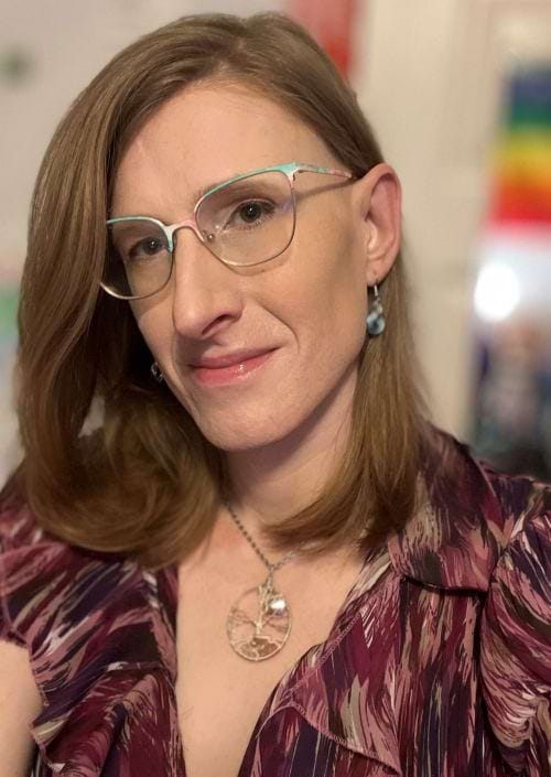 headshot of Emoria, a white trans woman in a purple ruffly blouse, a tree of life pendant, and trans-colored glasses, looking at the camera with head slightly tilted