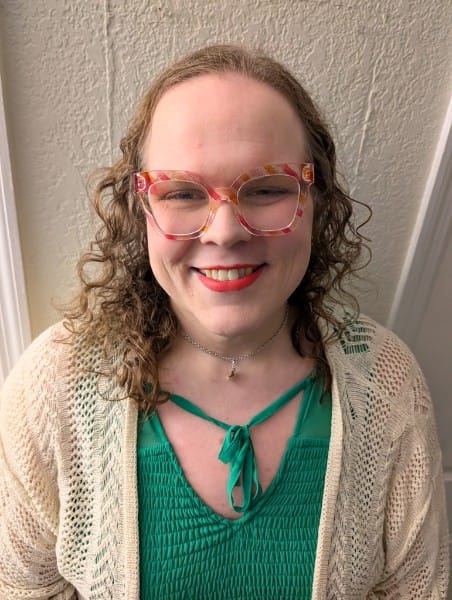 A smiling white trans girl in a green top, cream cardigan, and pop-art glasses framed in pink and orange stripes.