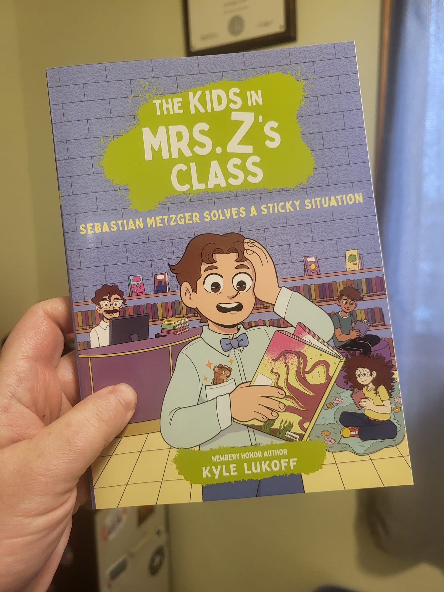 hand holding a copy of The Kids in Mrs Z's Class: Sebastian Metzger Solves a Sticky Situation. A drawing of a dismayed looking brown-haired boy with medium light skin holding a book covered with a stain, with a library scene behind him.