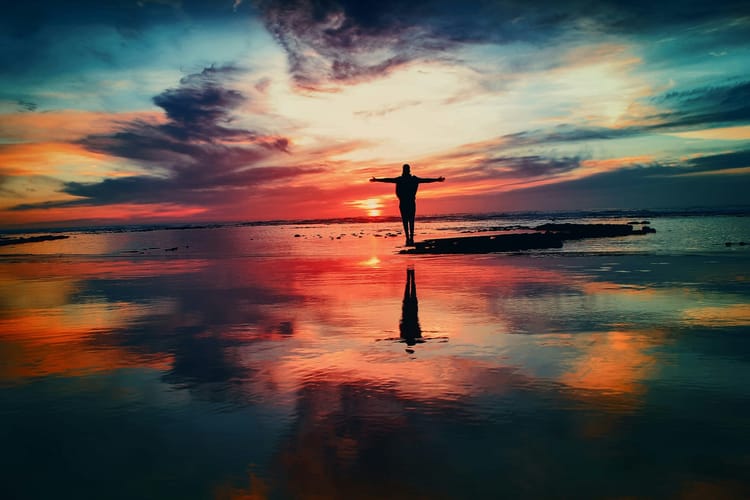 Silhouette of a person standing with arms outstretched amid a vibrant rainbow-colored sunset at the beach.